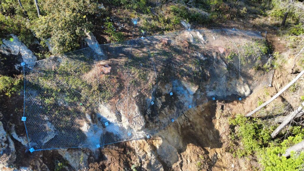 Filet métallique de protection contre les chutes de pierres sur une pente rocheuse boisée avec ancrages de sécurité.