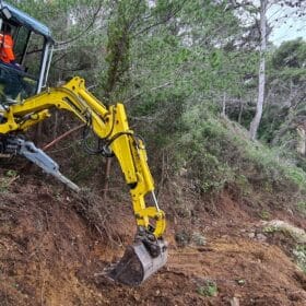 Pelle araignée jaune effectuant un terrassement délicat sur une pente boisée escarpée et boueuse.