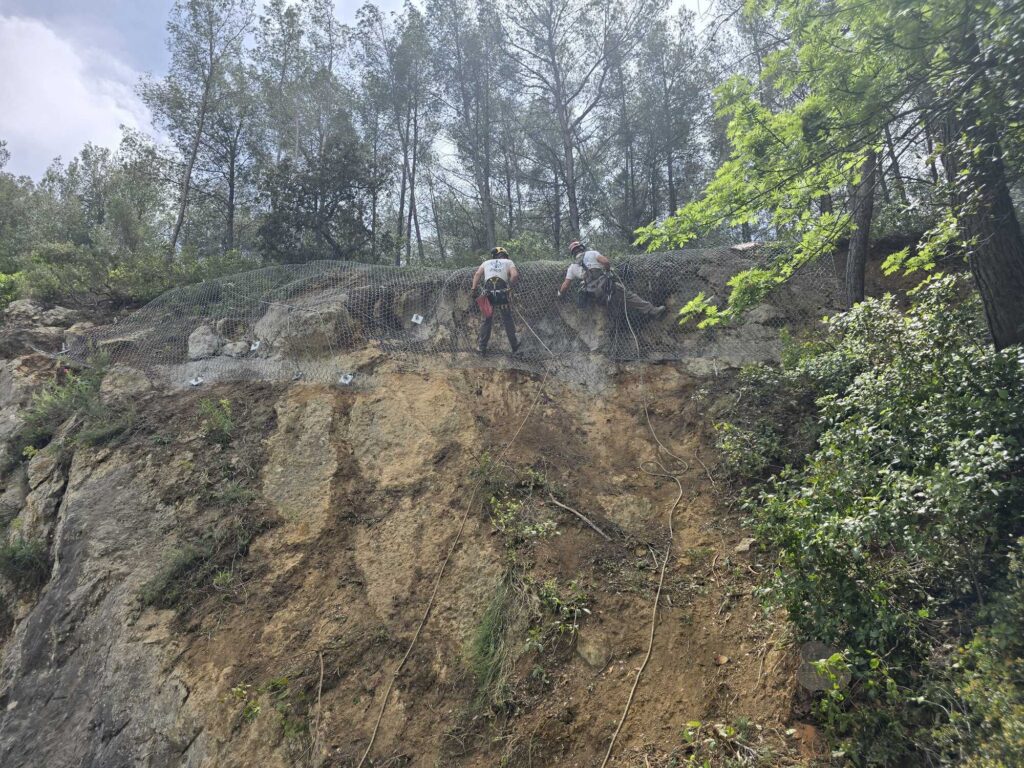 Techniciens fixant un filet de protection contre les chutes de pierres sur un talus rocheux escarpé en forêt.